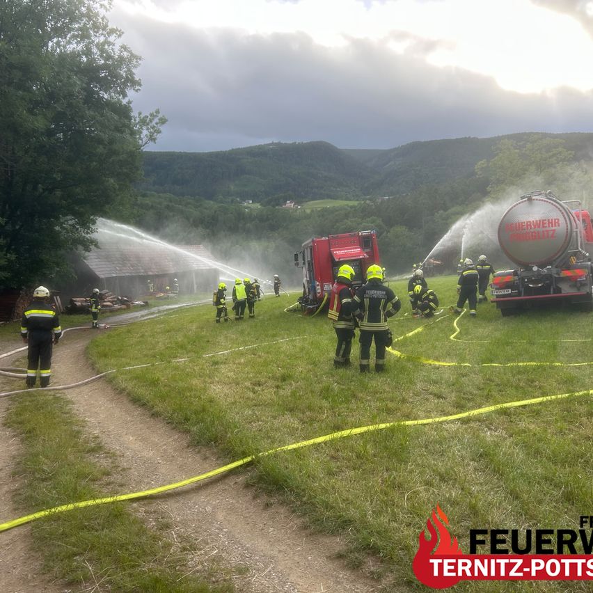 Feuerwehrleute löschen einen Brand in einer Scheune mit einem Tankwagen und einem Feuerwehrauto. Die Feuerwehrschläuche sind angeschlossen und die Feuerwehrleute tragen Schutzausrüstung.