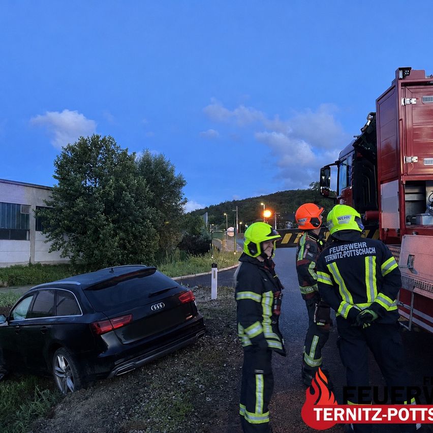 Drei Feuerwehrleute in Schutzkleidung stehen neben einem roten Feuerwehrauto. Ein schwarzer Audi ist am Straßenrand geparkt.
