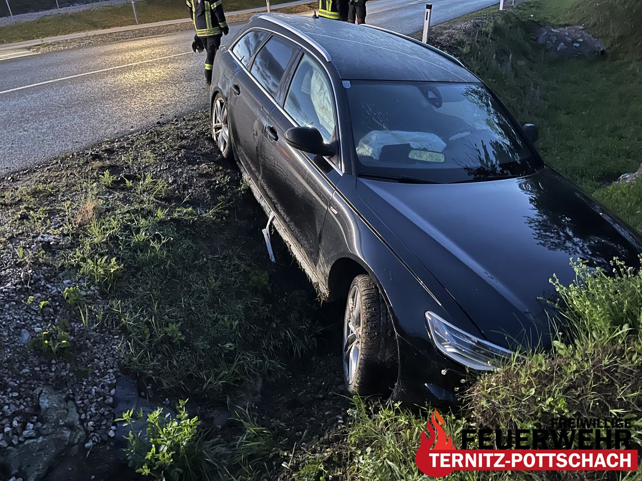 Ein schwarzer Wagen ist in einen Graben am Straßenrand gecrasht, mit anwesenden Rettungskräften. Das Auto hat vorne und hinten Schäden erlitten und die Fahrertür steht offen.