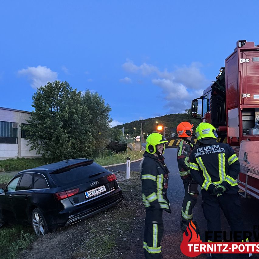 Ein Autounfall ereignete sich in der Nähe eines Feuerwehrwagens. Feuerwehrleute in Uniformen stehen neben dem Fahrzeug. Ein Gebäude und Bäume sind im Hintergrund.