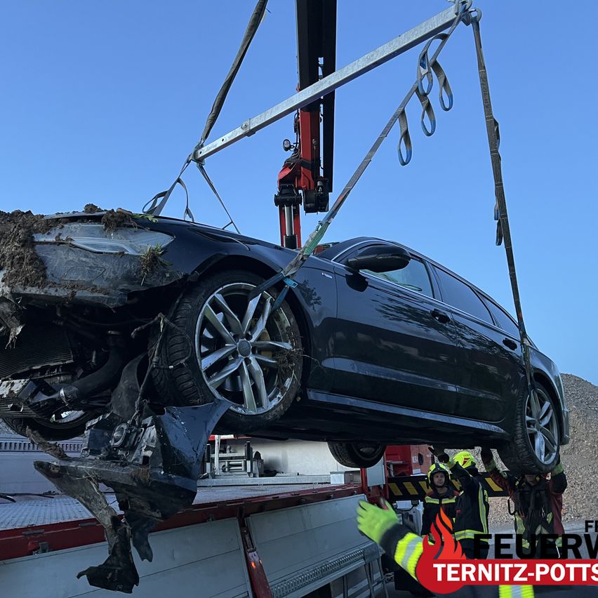 Ein beschädigtes schwarzes Auto wird von einem Kran angehoben. Feuerwehrleute arbeiten daran. Es ist ein sonniger Tag.