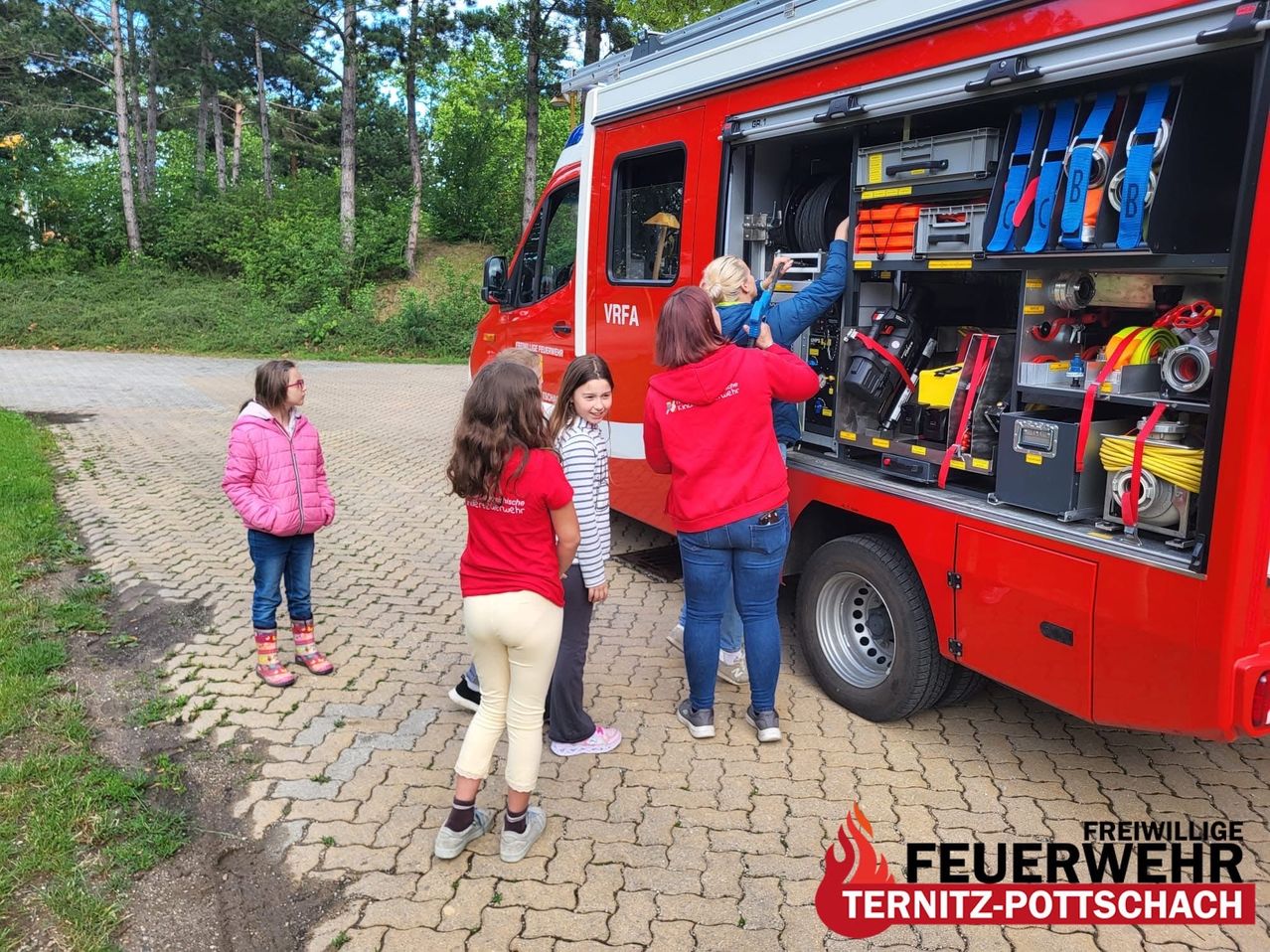 Ein Feuerwehrauto ist geöffnet, wobei mehrere Erwachsene und Kinder beobachten. Das Auto hat die Buchstaben VRFA an der Seite. Die Erwachsenen greifen nach Ausrüstung.
