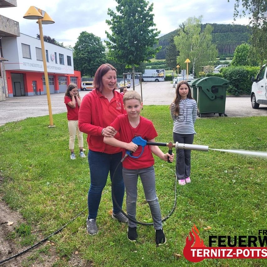 Eine Frau und ein Junge in Rot halten einen Wasserschlauch und sprühen Wasser. Dahinter schaut ein Mädchen zu. In der Nähe steht ein Feuerhydrant, ein Mülleimer und ein Gebäude.