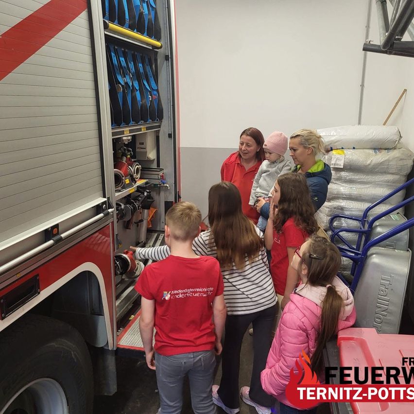 Eine Gruppe von Kindern und Erwachsenen inspiziert einen Feuerwehrwagen. Die Kinder tragen rote T-Shirts, und eine Frau hält ein Baby. Ausrüstung und Vorräte sind im Inneren des Wagens zu sehen.
