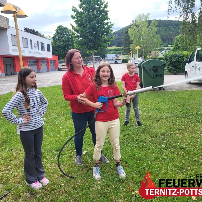 Eine Gruppe von Kindern und Erwachsenen übt das Benutzen eines Feuerlöschers auf Gras, mit einem Feuerwache im Hintergrund.