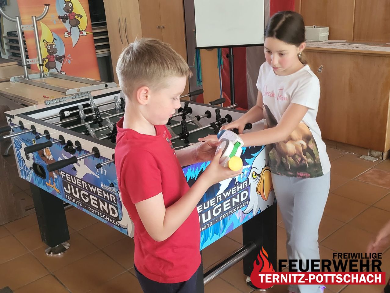Ein Junge und ein Mädchen spielen mit einem gelben Spielzeug in der Nähe eines Tischfußballs mit einem 'Feuerwehr Jugend'-Banner.