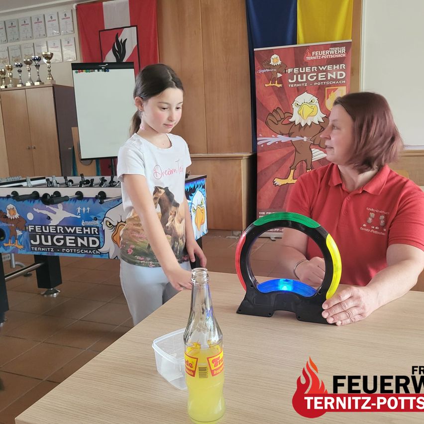 Ein junges Mädchen führt ein Experiment mit einer Flasche Flüssigkeit und einem farbigen Rad in einem Raum mit einem Feuerwehr-Poster durch.