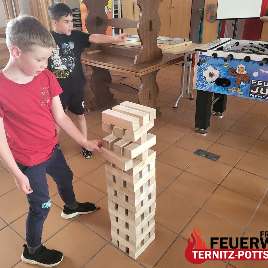 Zwei Jungen spielen mit einem hohen Stapel aus Holzklötzen, einer von ihnen trägt ein rotes T-Shirt. Im Hintergrund gibt es einen Tischkicker und einen Tisch.