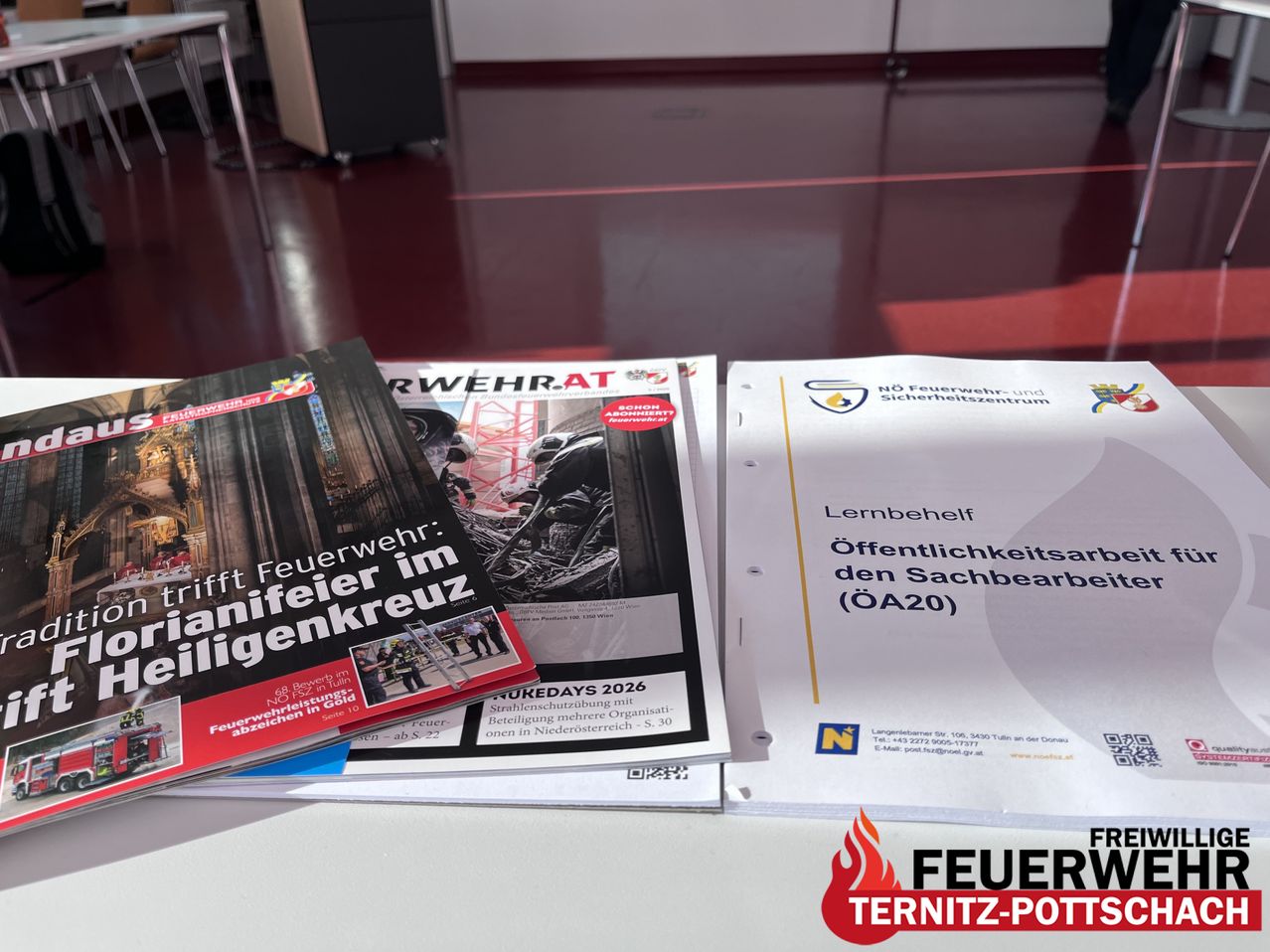 Magazine, Broschüren und Papiere liegen auf einem Tisch. Die oberste ist mit 'Feuerwehr:' beschriftet und zeigt Feuerwehrleute. Darunter ist 'Rwehr-AT.' Ein weiteres Papier trägt den Titel 'Lernbehelf' und hat ein Sicherheitslogo.