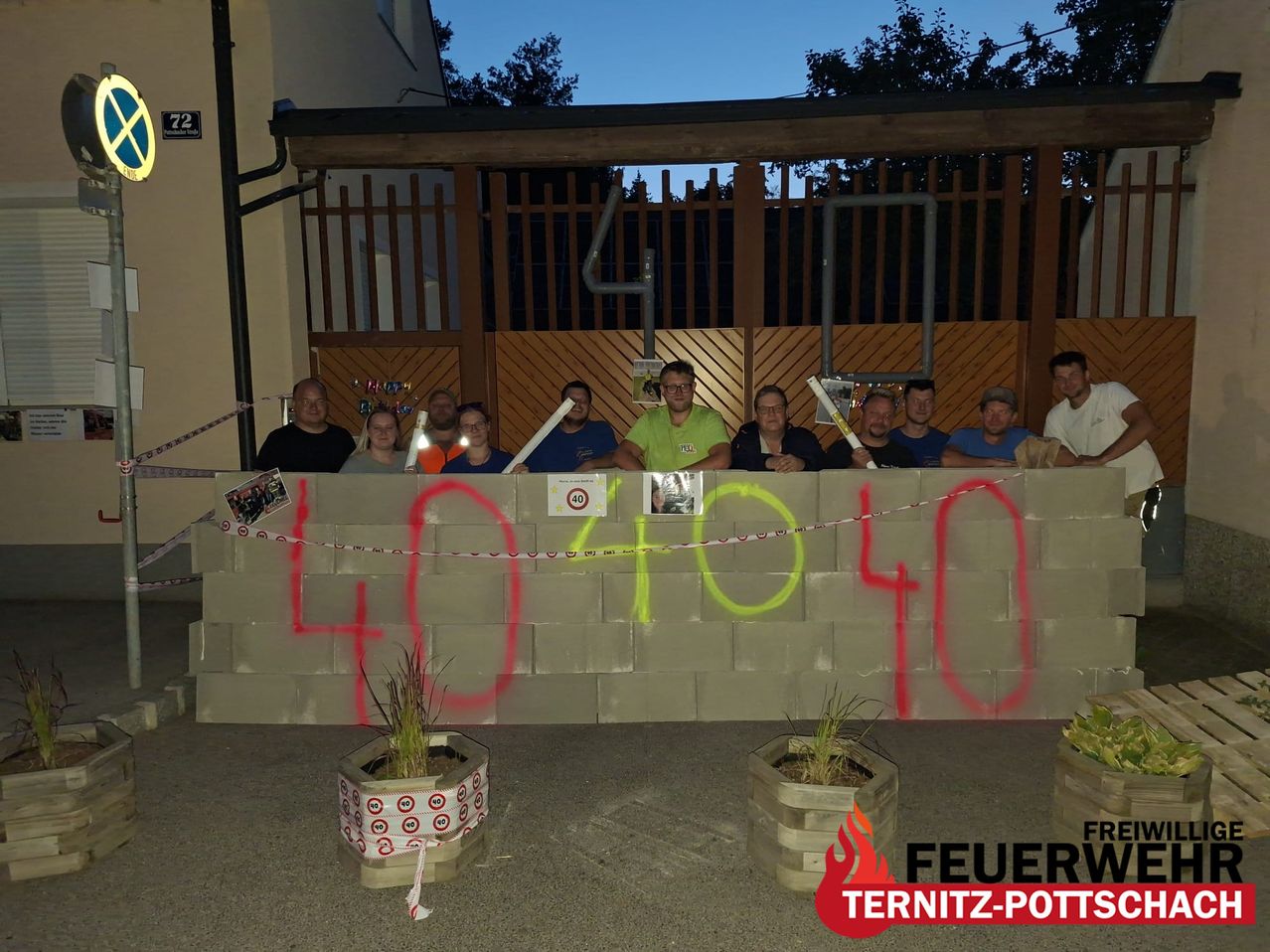 A group of people are standing behind a concrete wall with graffiti reading '404040'. They are holding sticks. There are potted plants on either side of the wall.