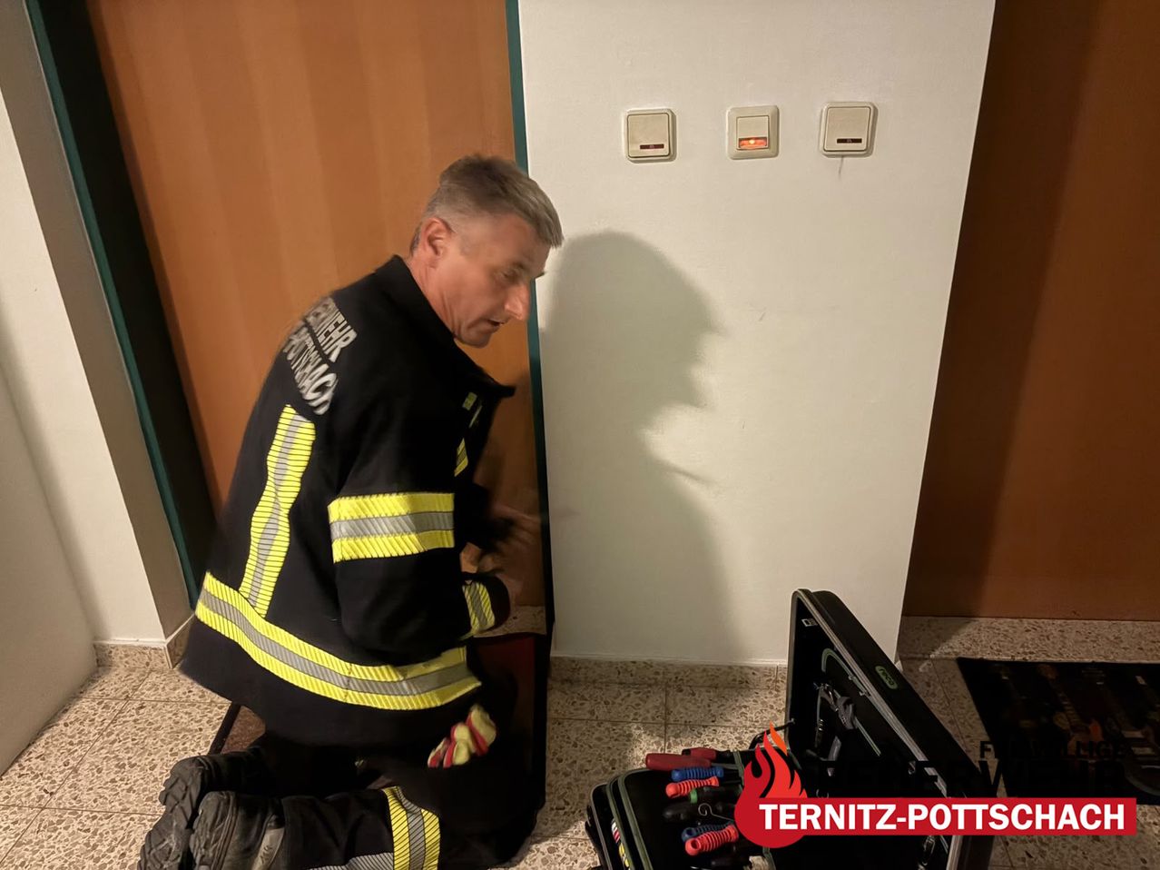 A firefighter is working on a door while kneeling on the floor, wearing a uniform. A toolbox with tools is open next to him.