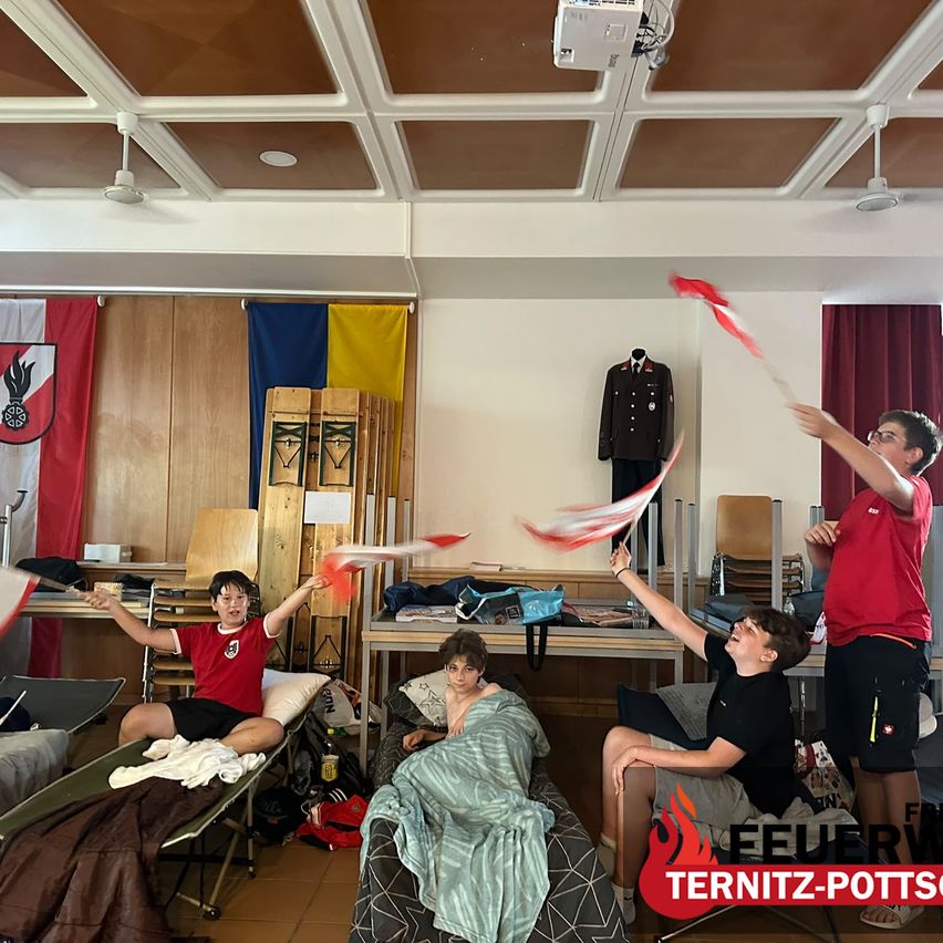 A group of boys in red shirts play with red flags in a room. A girl sits on a bed, and another boy holds a flag. There are flags, a mannequin, and a bag on the table.