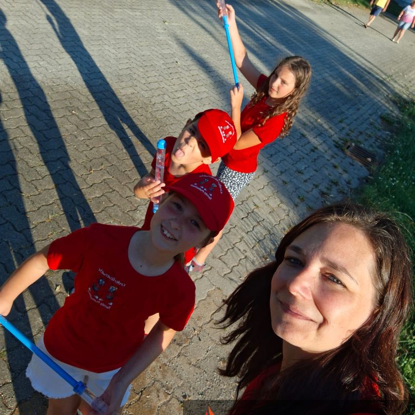 Three children and a woman are outside. The children are holding blue sticks, and the woman is smiling. They are on a brick floor with shadows and some people in the background.