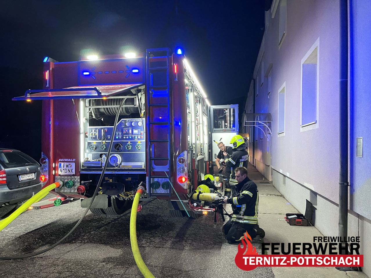 Firefighters in full gear are working on a red fire truck with a hose connected. They are standing in front of a building with a lit-up sign.