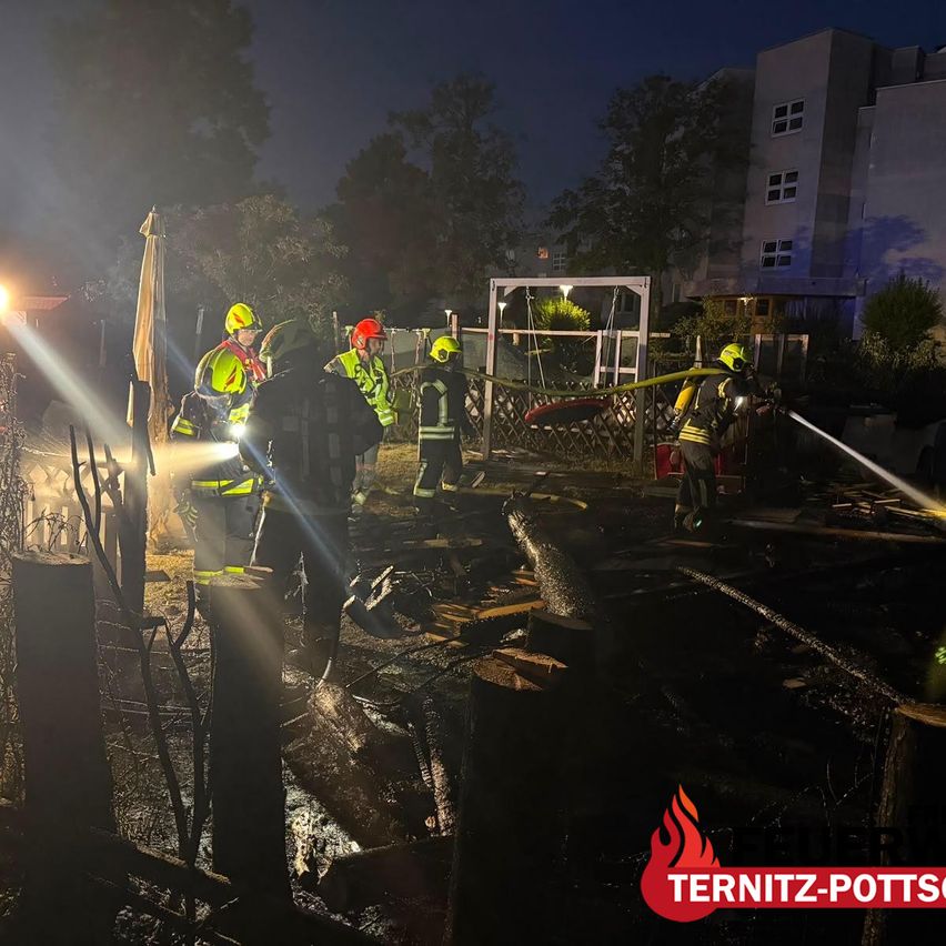 Firefighters in safety gear work at night in a burned-out area. A bright light illuminates the scene. The area is surrounded by a fence and trees.