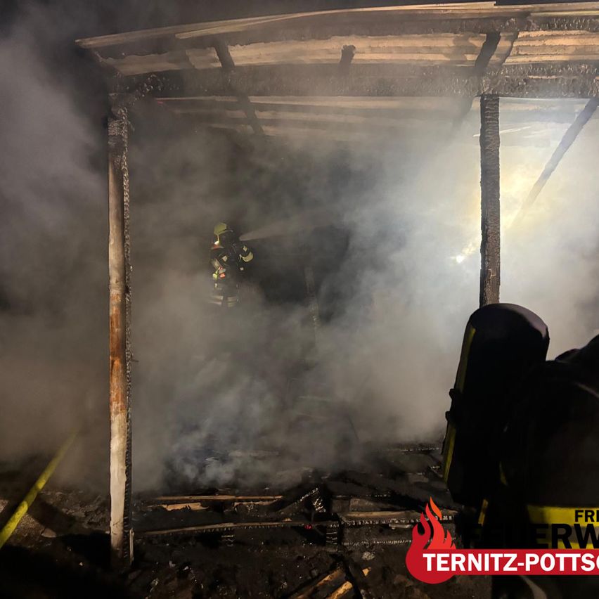 Firefighters are in a smoke-filled shed, wearing protective gear. The shed's roof is damaged, and smoke billows out. The ground is littered with debris.