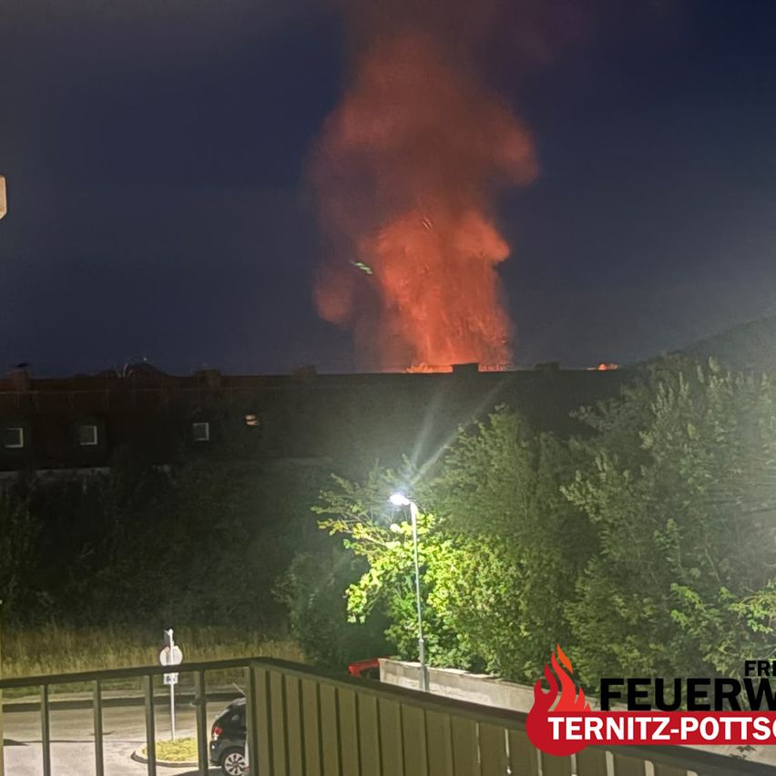 A large fire at night, with a burning building and smoke rising. Trees and street lights are visible in the foreground.