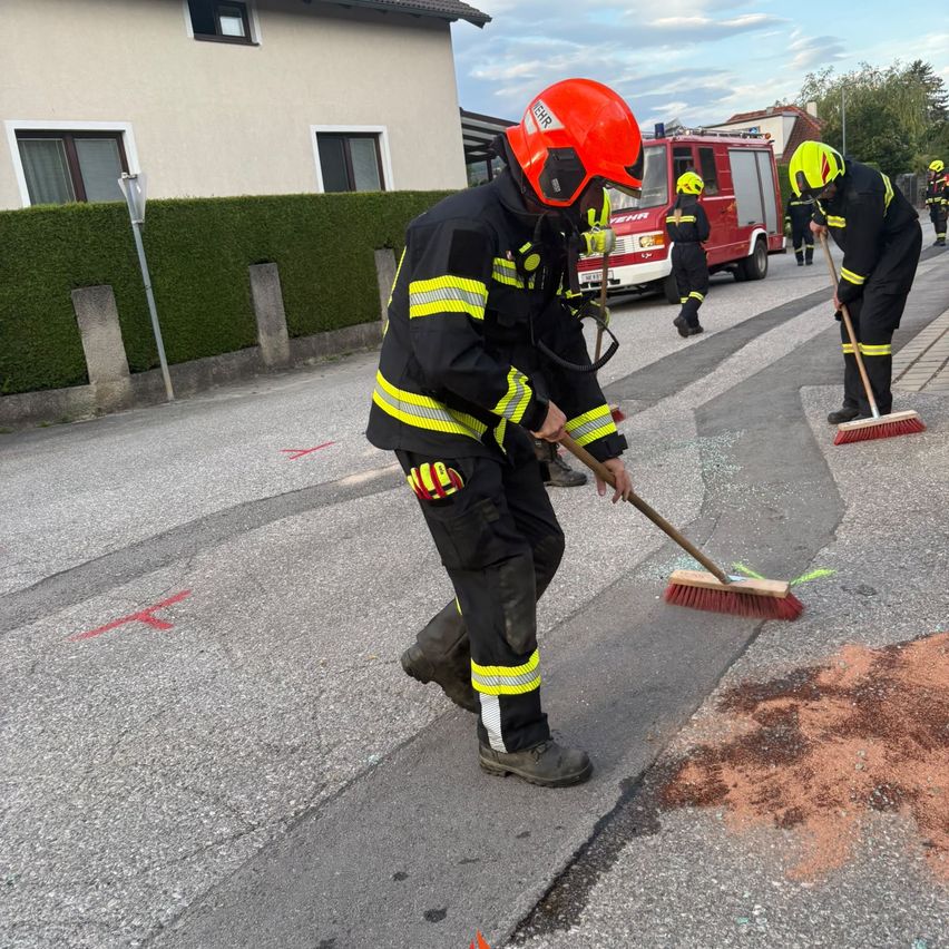 Firefighters in protective gear clean up a spill on the street using brooms. A red truck is parked nearby.