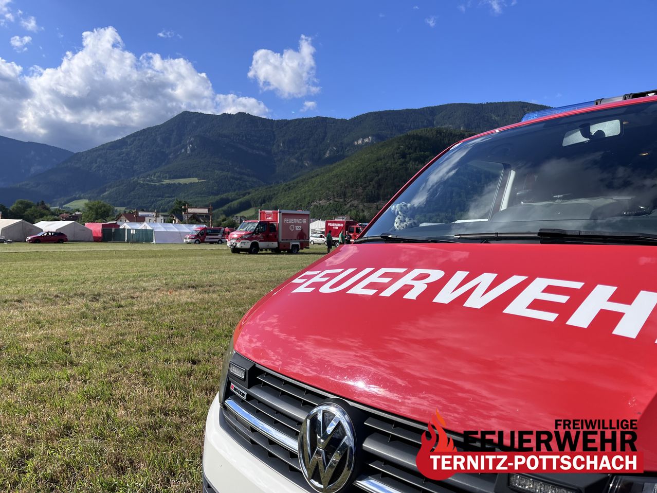 A red VW fire truck parked on a grassy field with mountains in the background. Several tents and vehicles are in the area.