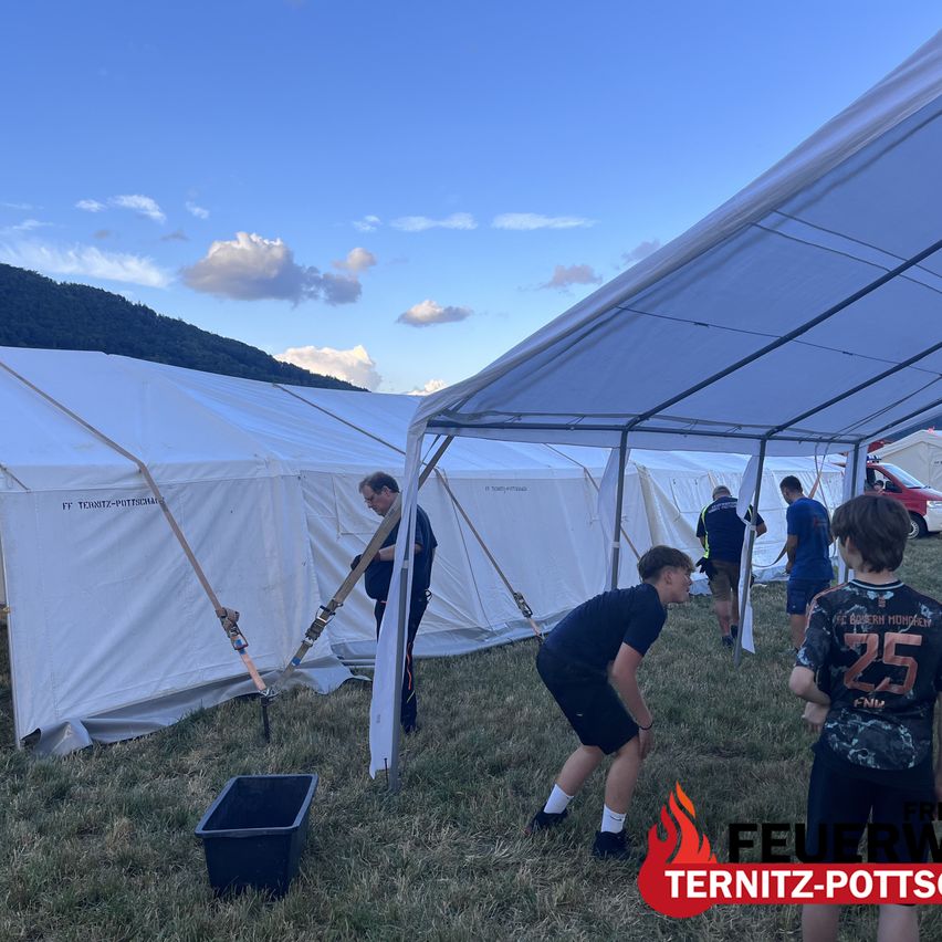 Several people are setting up tents on a grassy field under a clear blue sky. One man is adjusting the tent poles while others look on. A black storage box is nearby.