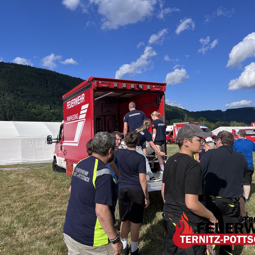 A group of people are gathered around a red fire truck, with some standing near the open back door. A few people are inside the truck. The scene takes place outdoors with a clear blue sky and mountains in the background.