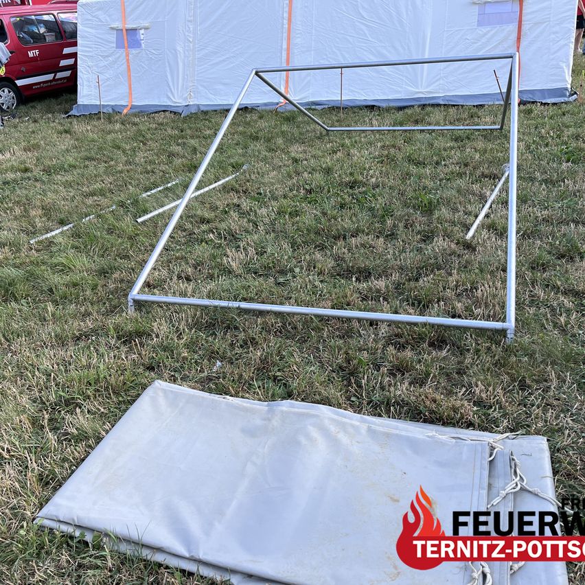 A metal frame structure is on a grassy field with a tent in the background. A gray cover is laid out nearby. A watermark reads 'FEUER'.