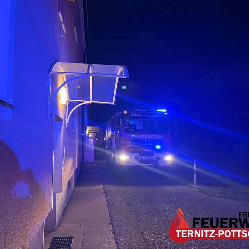 A fire truck with flashing lights is parked in front of a building at night. The building has a blue wall and a glass canopy. The image is taken from a low angle.