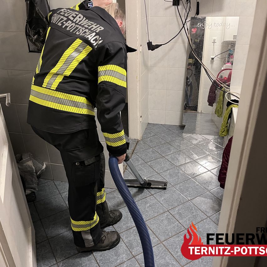 A firefighter in full gear is using a hose to clean a tiled bathroom floor. The room is small with white tiles and a mirror on the wall.