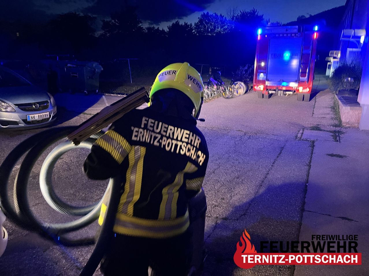 A firefighter in a yellow helmet is holding a hose, with a fire truck in the background at night.