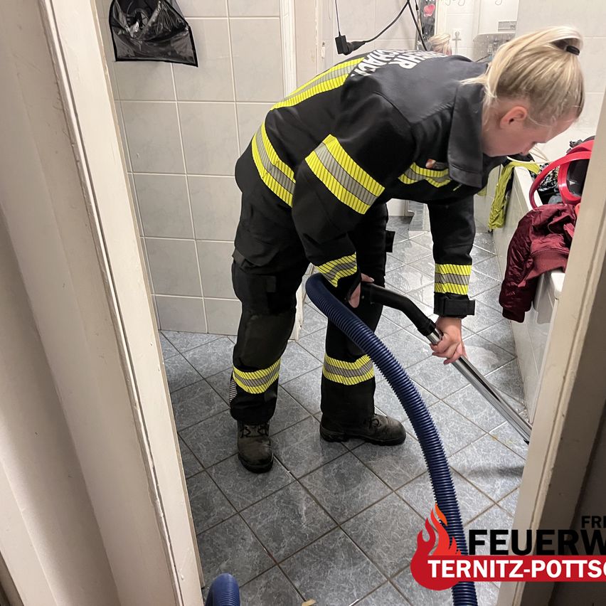 A woman in a firefighter uniform is cleaning the floor with a vacuum in a bathroom. The floor is tiled, and there is a black bag hanging on the wall.