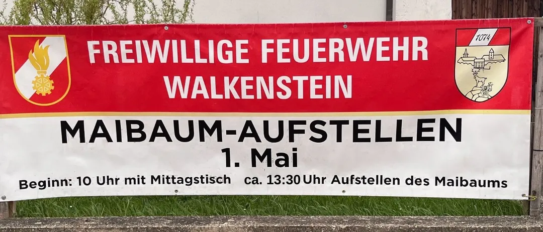 Ein roter und weißer Banner für die Freiwillige Feuerwehr Walkenstein ist an einer Steinmauer befestigt. Der Banner steht 'Maibaum-Aufstellen' und gibt an, dass die Veranstaltung um 10 Uhr mit Mittagspause um 13:30 Uhr beginnt.