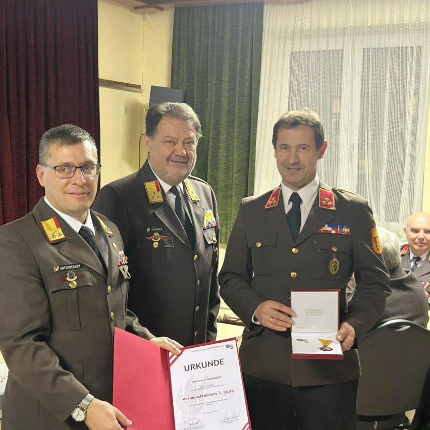 Drei Männer in Militäruniformen stehen nebeneinander und lächeln. Der Mann auf der linken Seite hält eine rote Mappe mit einem Zertifikat darin, während der Mann auf der rechten Seite eine Medaille in einer Box hält. Dahinter ist ein verschwommener Hintergrund mit einem Vorhang und einem Fenster zu sehen.