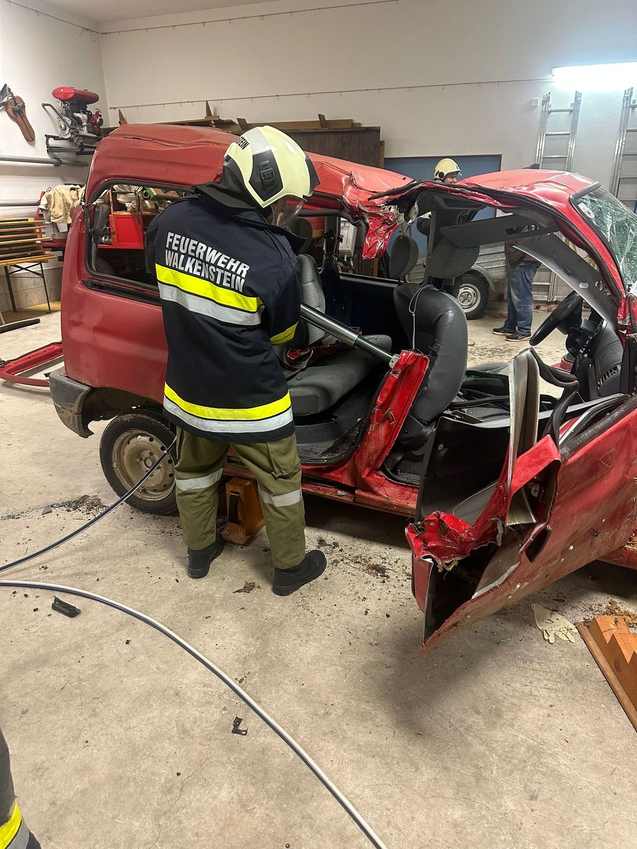 Ein Feuerwehrmann in einer roten Uniform mit 'Feuerwehr Walkenstein' auf dem Rücken inspiziert ein beschädigtes rotes Fahrzeug in einer Garage.