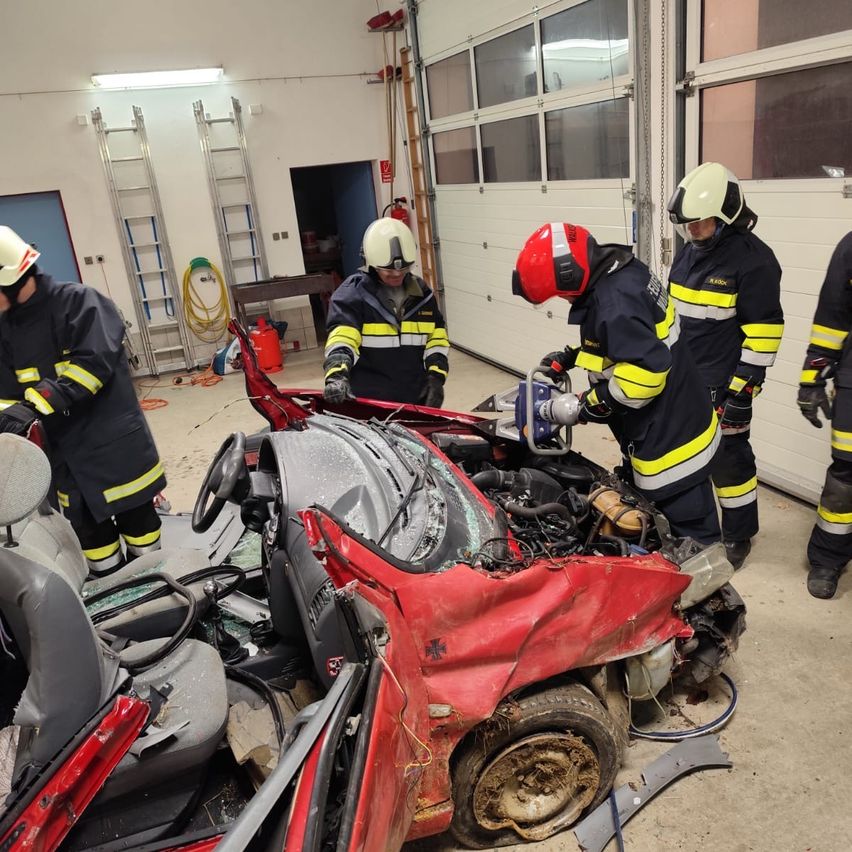 Eine Gruppe von Feuerwehrleuten in Uniform inspiziert ein beschädigtes rotes Auto in einer Garage. Das Auto hat eine zerbrochene Windschutzscheibe und einen freiliegenden Motor. Zwei Feuerwehrleute verwenden Werkzeuge am Auto.