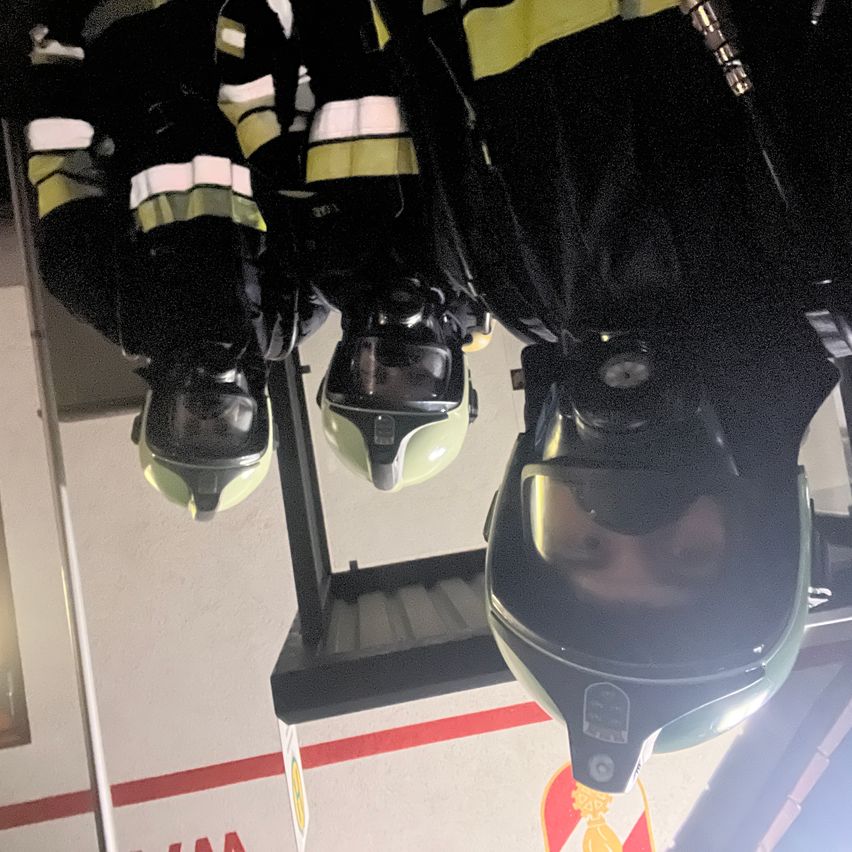 Drei Feuerwehrleute in Schutzausrüstung stehen in einem verspiegelten Flur und tragen Helme. Die Helme reflektieren die Umgebung und einander und erzeugen ein vielschichtiges Bild.