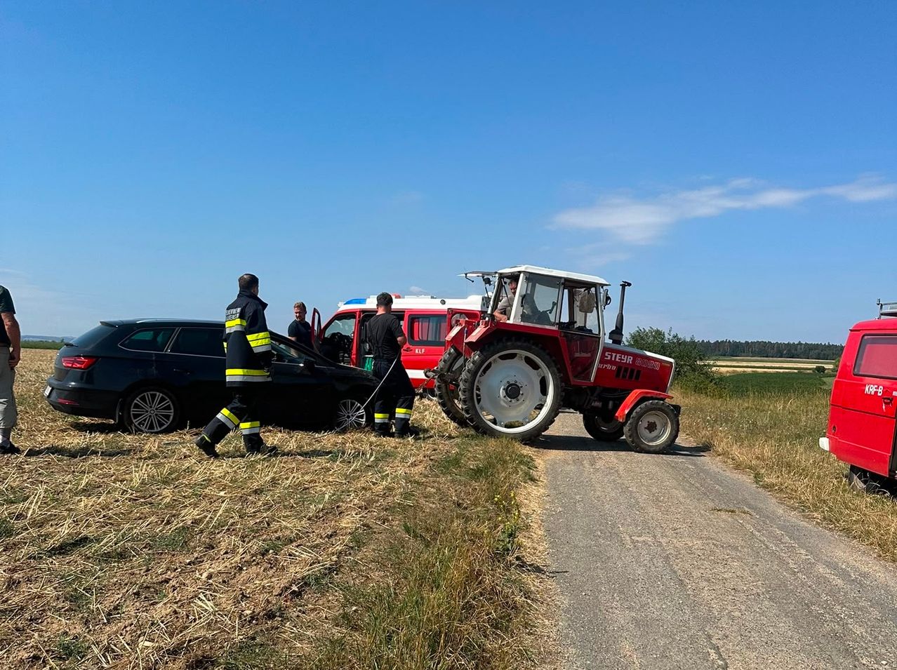 Rettungskräfte und ein Traktor stehen neben einem beschädigten Auto auf einer Landstraße unter klarem Himmel.