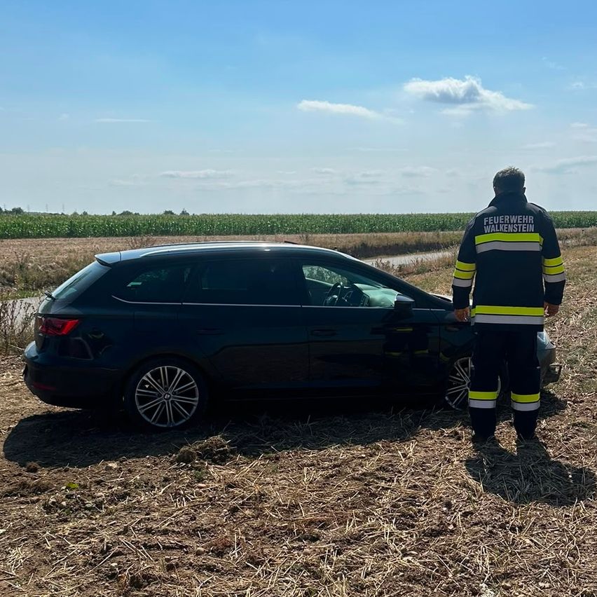 Ein Feuerwehrmann steht neben einem schwarzen Auto, das in ein Feld gecrasht ist. Das Auto steht auf dem Boden, und der Feuerwehrmann trägt eine reflektierende Sicherheitsweste.