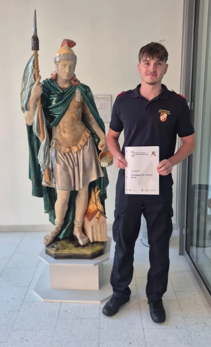 Ein Mann in Uniform steht neben einer Statue eines römischen Soldaten, der einen Schild und einen Becher hält, mit einem feuergeschädigten Haus im Hintergrund.