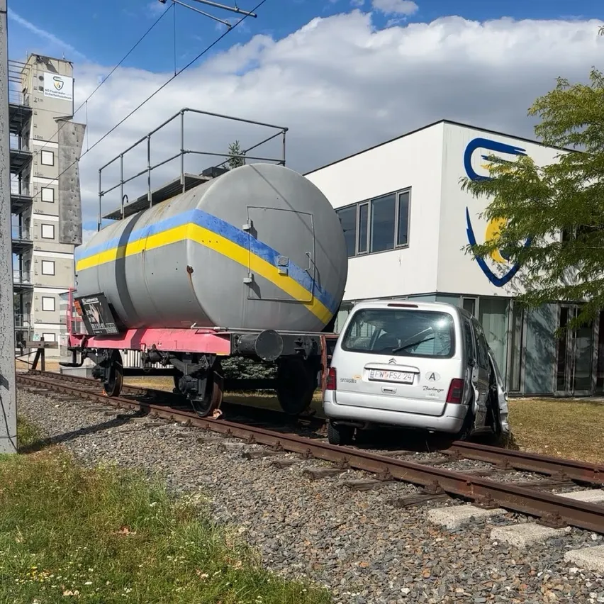 Ein silberner Van ist auf einem Bahngleis mit einem beschädigten Frontteil geparkt, neben einem großen Tankwagen mit gelben und blauen Streifen.