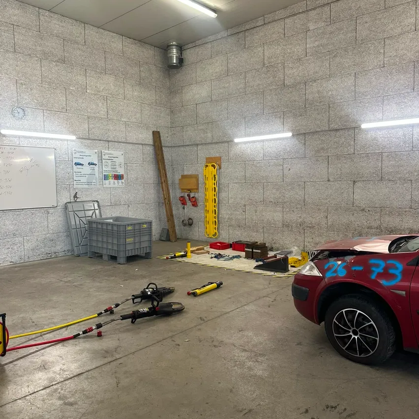 Eine überdachte Garage mit einem roten Auto, Werkzeugen und Ausrüstung auf dem Boden. Eine Tafel und Lagerboxen sind links, und ein beschädigtes Auto ist rechts.