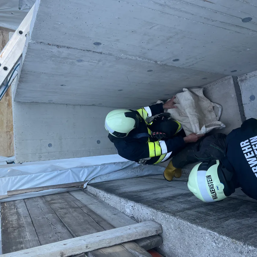 Zwei Feuerwehrleute in voller Ausrüstung sind in einer Betonstruktur. Einer hält einen Sack, während der andere auf den Stufen liegt.