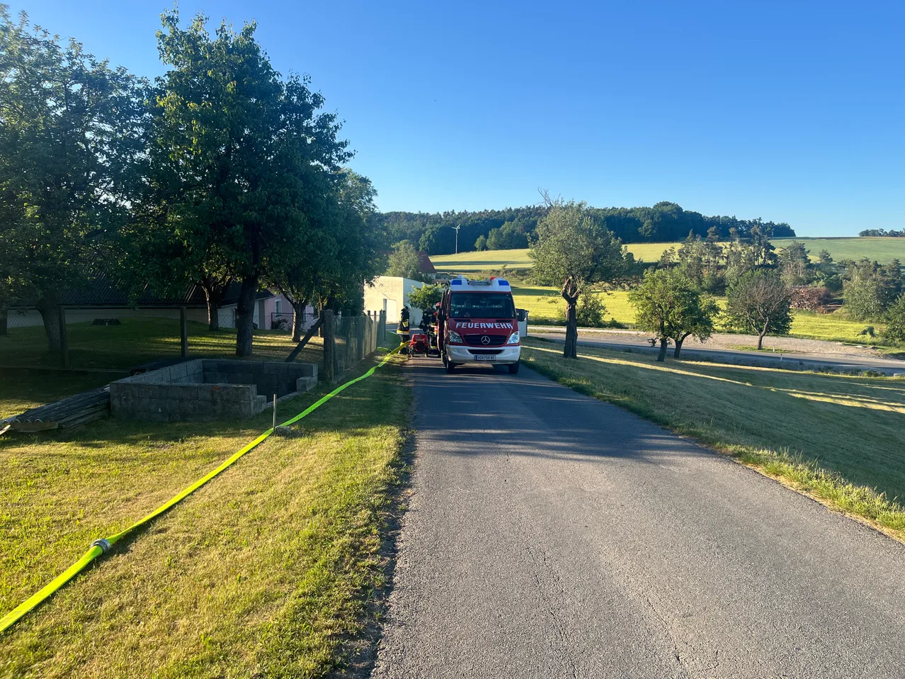 Ein Feuerwehrauto ist auf einer Landstraße geparkt, mit Bäumen auf beiden Seiten. Ein gelbes Absperrband liegt auf dem Boden. Ein Haus und ein Baum sind im Hintergrund zu sehen.