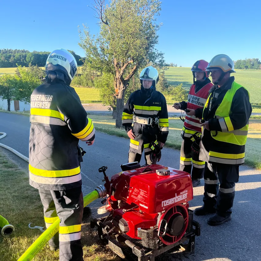 Feuerwehrleute stehen um einen roten Rosenbauer-Generator herum, der auf einer Straße mit Bäumen und grünem Gras in der Nähe steht. Sie sind in Schutzausrüstung gekleidet.
