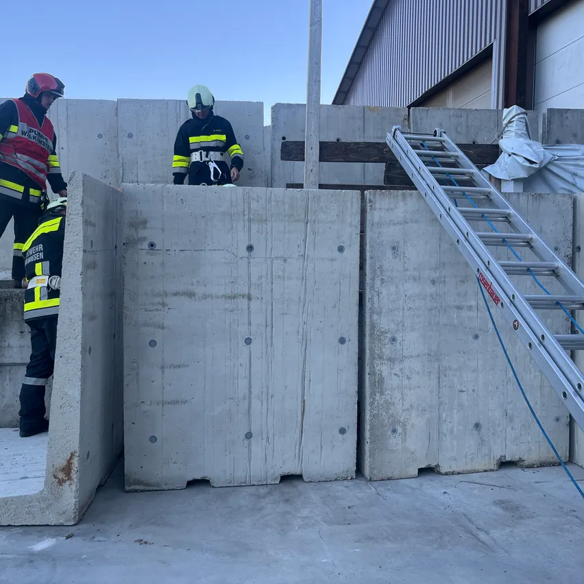 Drei Feuerwehrleute in Uniform stehen neben Betonbarrieren, an denen eine Leiter angelehnt ist. Dahinter befindet sich ein Gebäude.