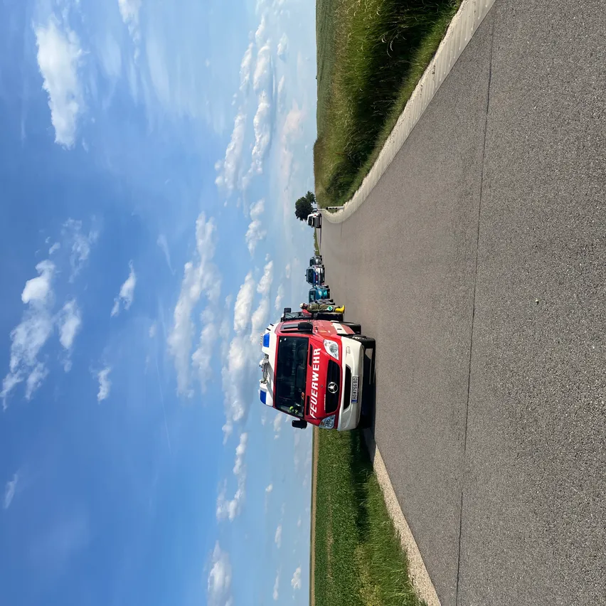Ein Feuerwehrauto fährt auf einer Landstraße mit mehreren anderen Fahrzeugen. Der Himmel ist blau mit verstreuten Wolken.