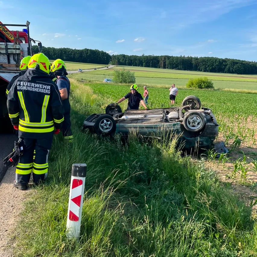 Feuerwehrleute untersuchen ein umgestürztes Auto am Straßenrand. Einige Leute stehen in der Nähe, und ein Feuerwehrauto ist geparkt.