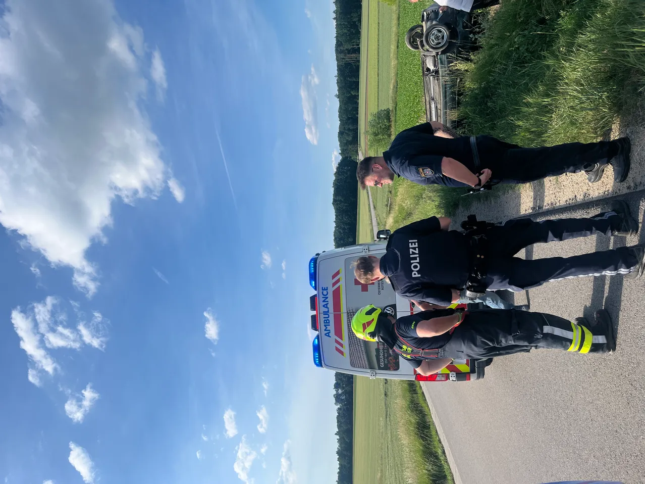 Polizisten stehen neben einem Krankenwagen. Ein umgekipptes Fahrzeug liegt im Gras. Der Himmel ist teilweise bewölkt.