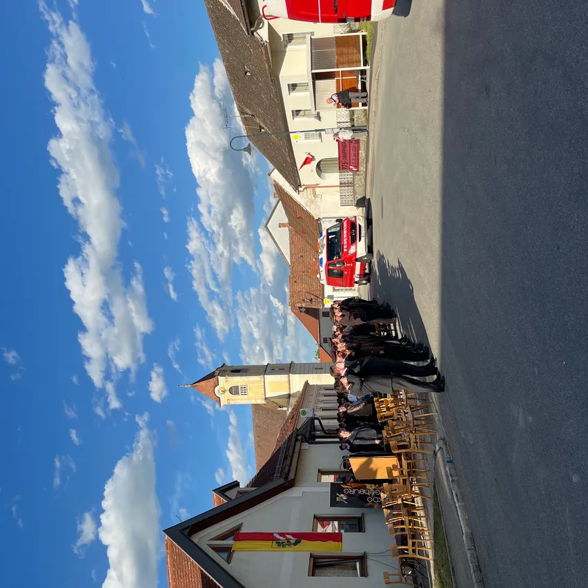 Eine Gruppe von Menschen steht in einer Reihe, vielleicht in Erwartung einer Veranstaltung. Sie sind in der Nähe eines Gebäudes mit Kirchturm und Fahne. In der Nähe ist ein rotes Rettungsfahrzeug geparkt. Der Himmel ist blau mit verstreuten Wolken.