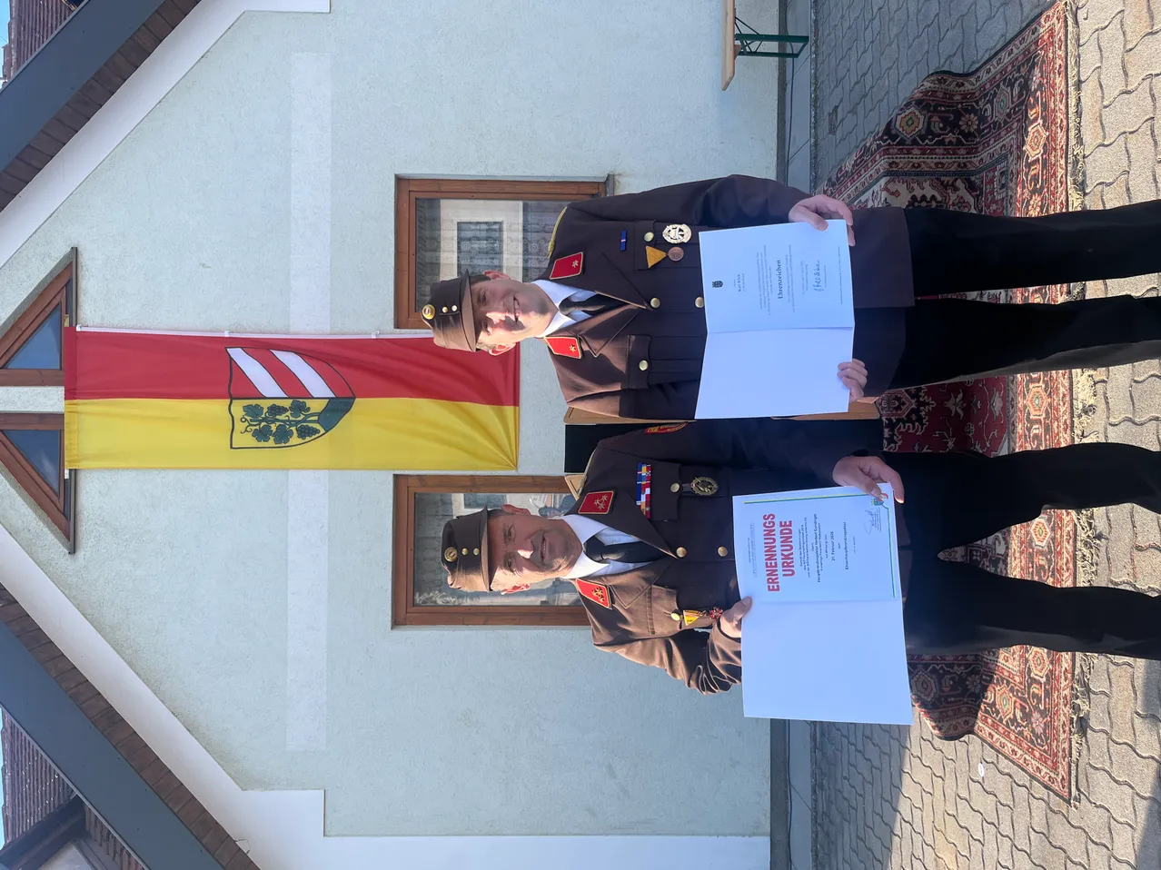 Zwei Militäroffiziere stehen vor einem Gebäude mit Urkunden in der Hand, einer hat eine Fahne mit Logo hinter ihnen.
