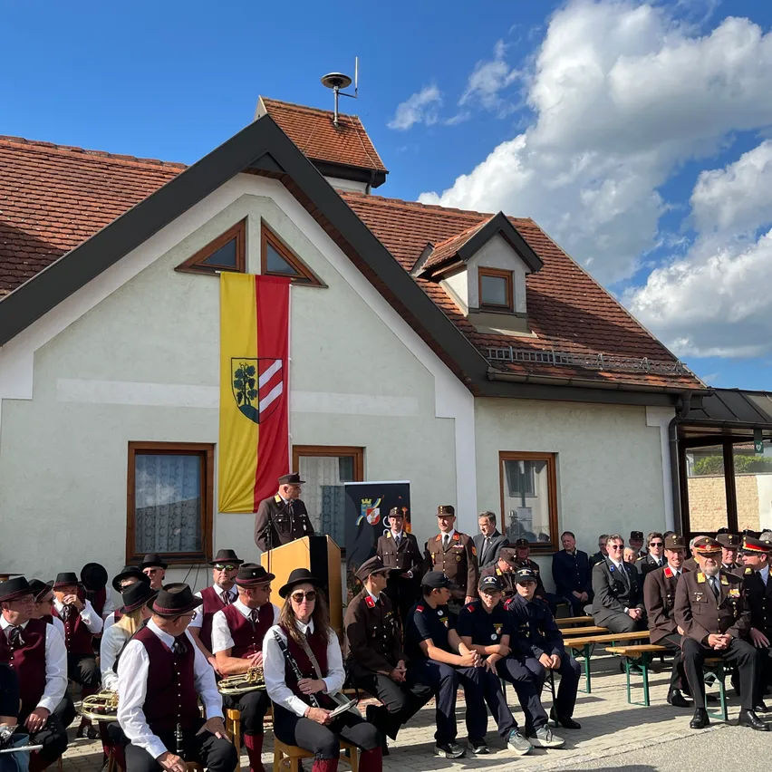 Eine Gruppe uniformierter Personen, darunter Musiker, versammelt sich vor einem Gebäude mit einer Fahne, wahrscheinlich für eine Zeremonie unter einem bewölkten Himmel.